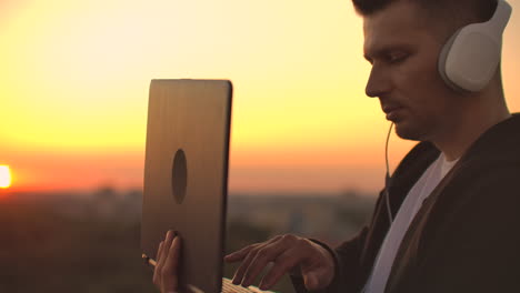 a man freelancer in headphones standing on the roof at sunset writes on the keyboard code pages. little business. listen to music and work at the computer enjoying the beautiful view from the roof.