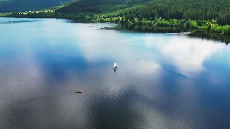 Un-Velero-En-Un-Lago-Sereno-En-La-Selva-Negra,-Alemania,-Rodeado-De-Exuberante-Vegetación,-Reflejando-Nubes,-Vista-Aérea