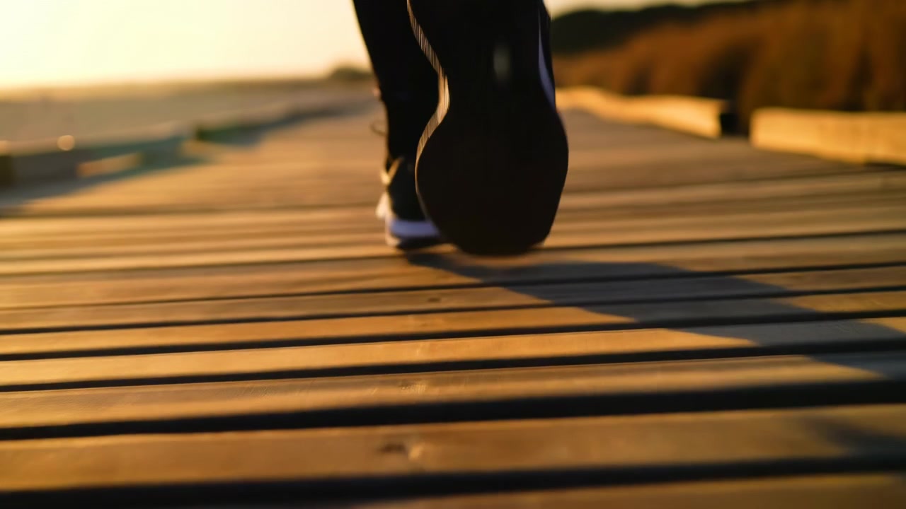 Prospettiva a livello del suolo mostra il tallone di una scarpa sportiva sollevato e la suola opposta che tocca una tavola di legno stagionata su una passerella costiera, con luce solare calda, texture, equilibrio e movimento