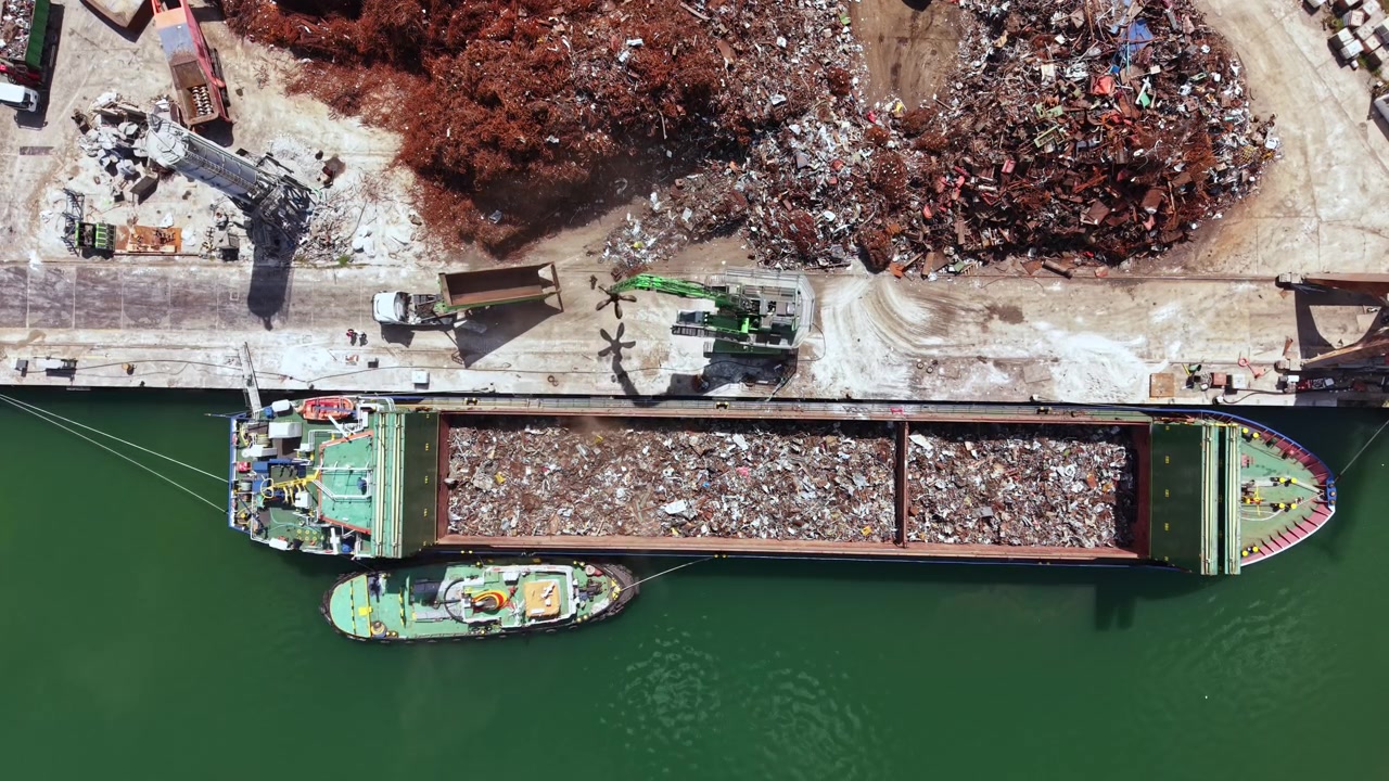 Un buque de carga descarga chatarra en un puerto cerca de un centro de reciclaje a mediodía con cielos despejados