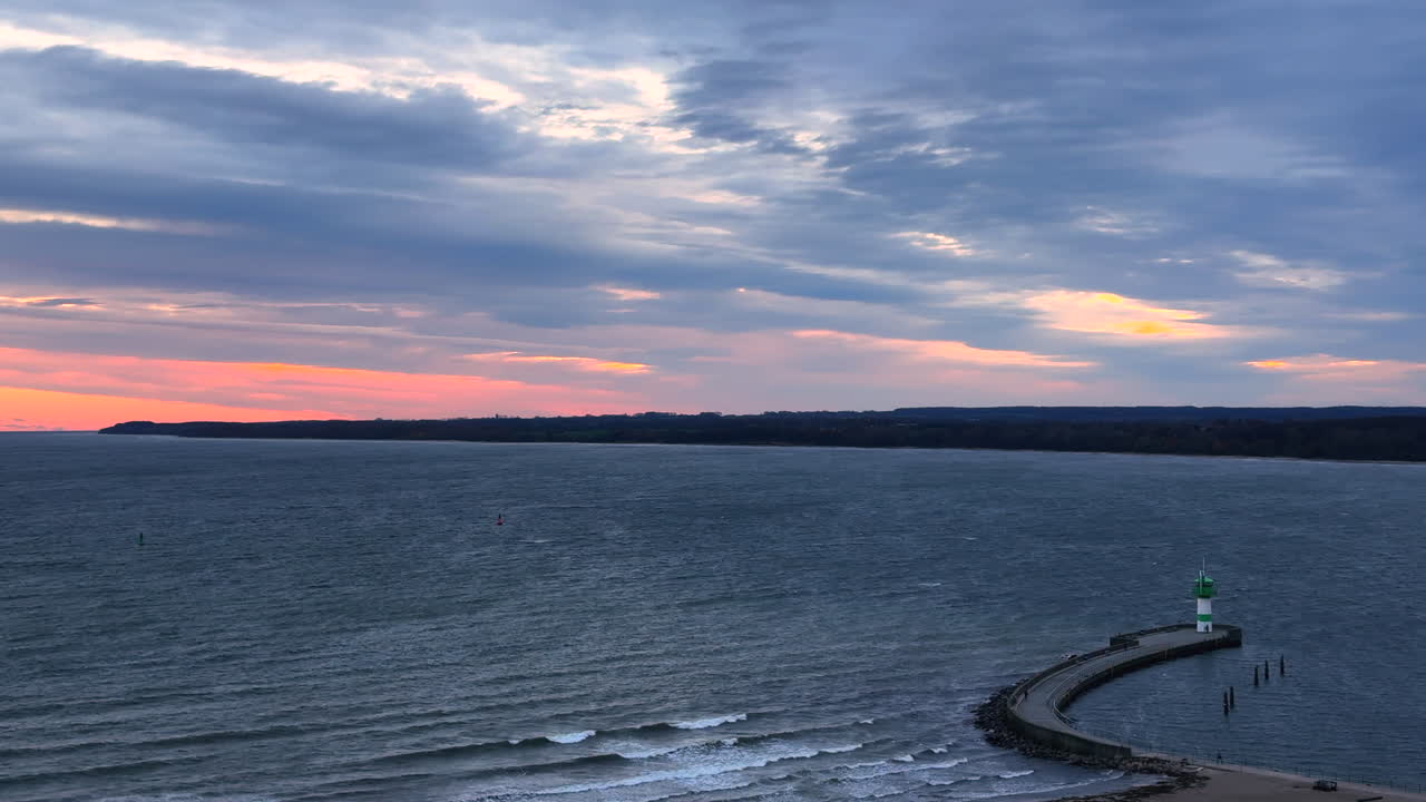 The video appears to be filmed in Travemünde and shows a windy coastal scene at the harbor entrance. A curved pier leads out to the green-and-white lighthouse at the breakwater