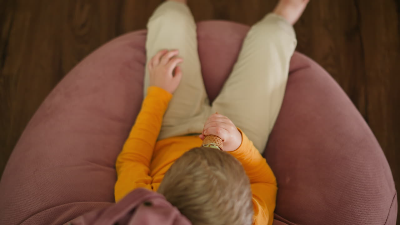 Aerial top view of young boy in orange top seated barefoot on pink beanbag, holding ice cream cone and casually playing with legs while relaxing indoors on wooden floor