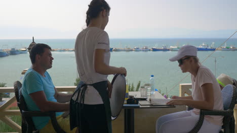 Young-couple-sitting-in-a-cafe-the-waitress-takes-the-order-from-the-woman-the-man-studying-the-menu