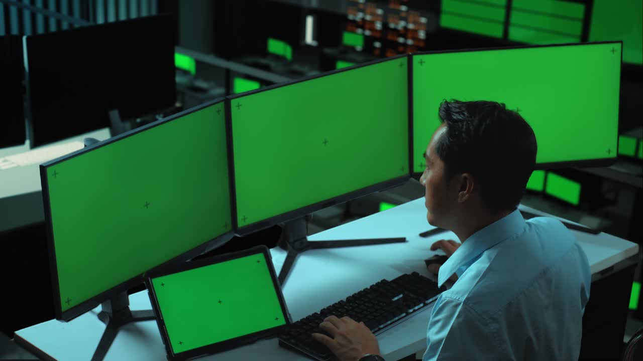 vista lateral de un hombre asiático trabajando con una tableta y varios monitores de computadora con una pantalla verde simulada en la oficina