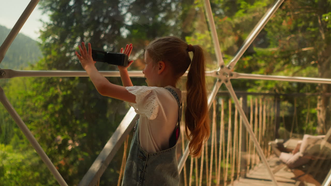 niño dispara en el lago del bosque en glamping. niña pequeña concentrada toma video de bosque salvaje en un hotel ecológico. niño con cámara digital en vacaciones