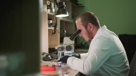 una vista de cerca de un técnico trabajando meticulosamente en un componente electrónico bajo un microscopio en un laboratorio, el espacio de trabajo está equipado con una lámpara de escritorio y varias herramientas