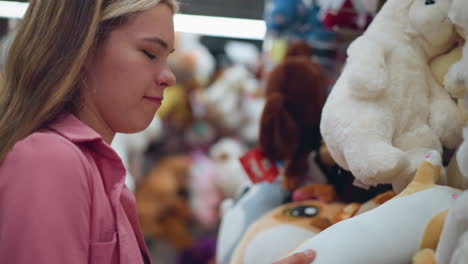 lady in pink dress stands in a brightly lit toy store, holding a plush toy, she playfully touches and inspects the texture of the toy with a smile on her face, surrounded by other colorful toy