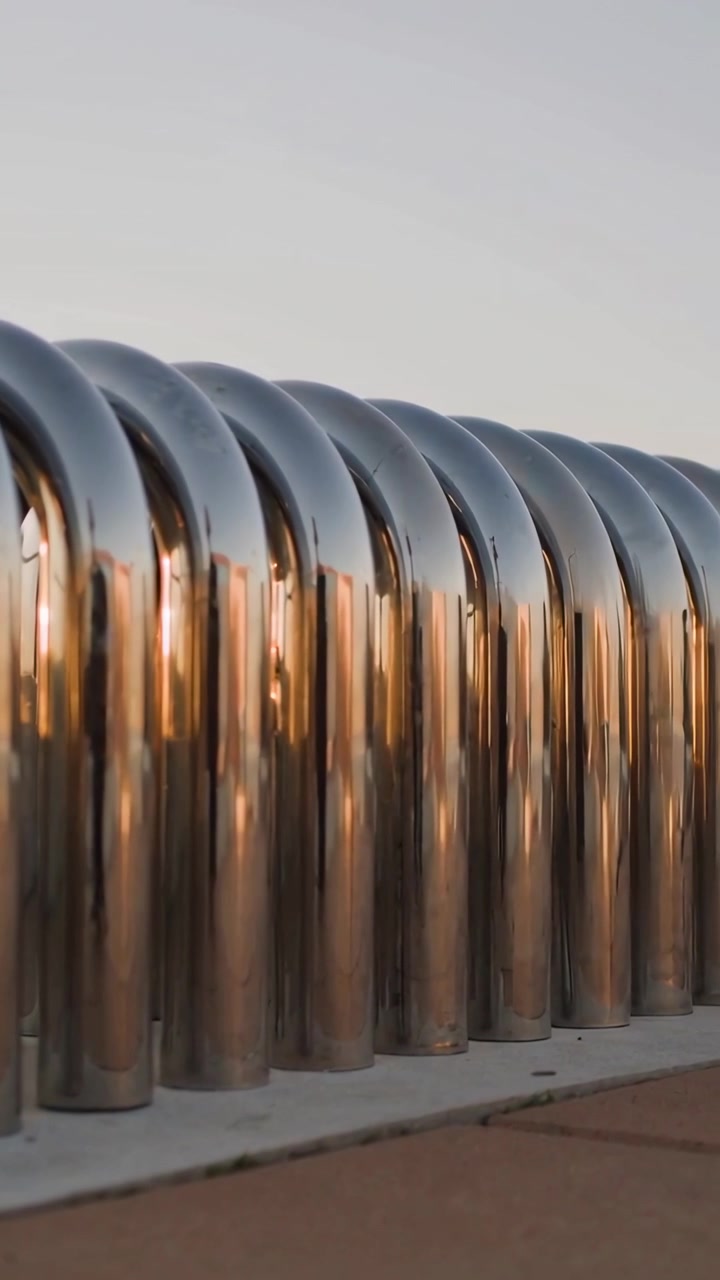 Vertical video: Panning camera revealing chrome arches of sculpture at plaza, shifting sunset light