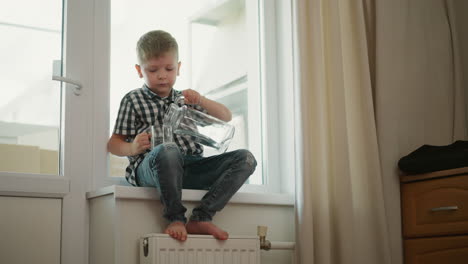 kid thoughtfully seated near wardrobe pouring clear water from large glass jar into transparent cup while resting on windowsill in casual clothes with natural daylight