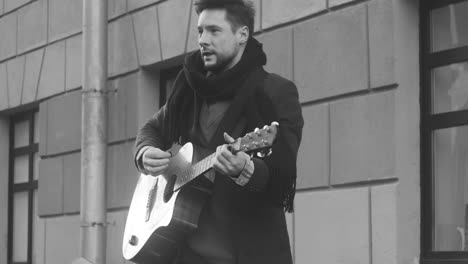 Vista-En-Blanco-Y-Negro-De-Un-Hombre-Con-Abrigo-Cantando-Y-Tocando-La-Guitarra-En-La-Calle