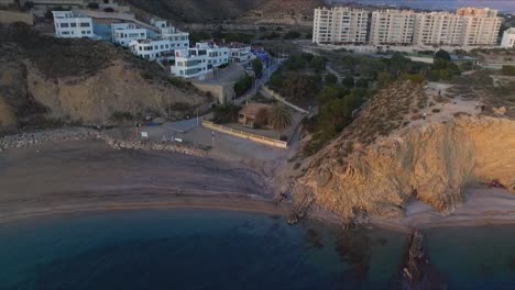 Die-Luftaufnahme-Erfasst-Die-Weißen-Gebäude,-Wohnblocks,-Eigentumswohnungen-Und-Die-Dramatische-Felswand-Am-Strand-Von-Es-Bol-Nou-Und-Unterstreicht-Die-Mischung-Aus-Architektur-Und-Natürlicher-Schönheit