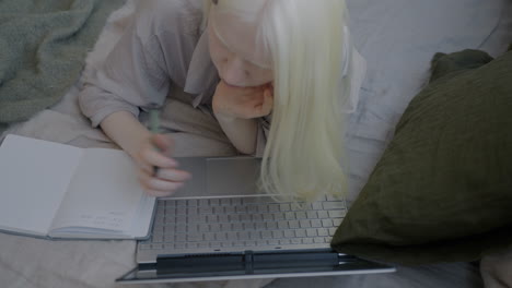 Young woman working from bed with laptop and notebook