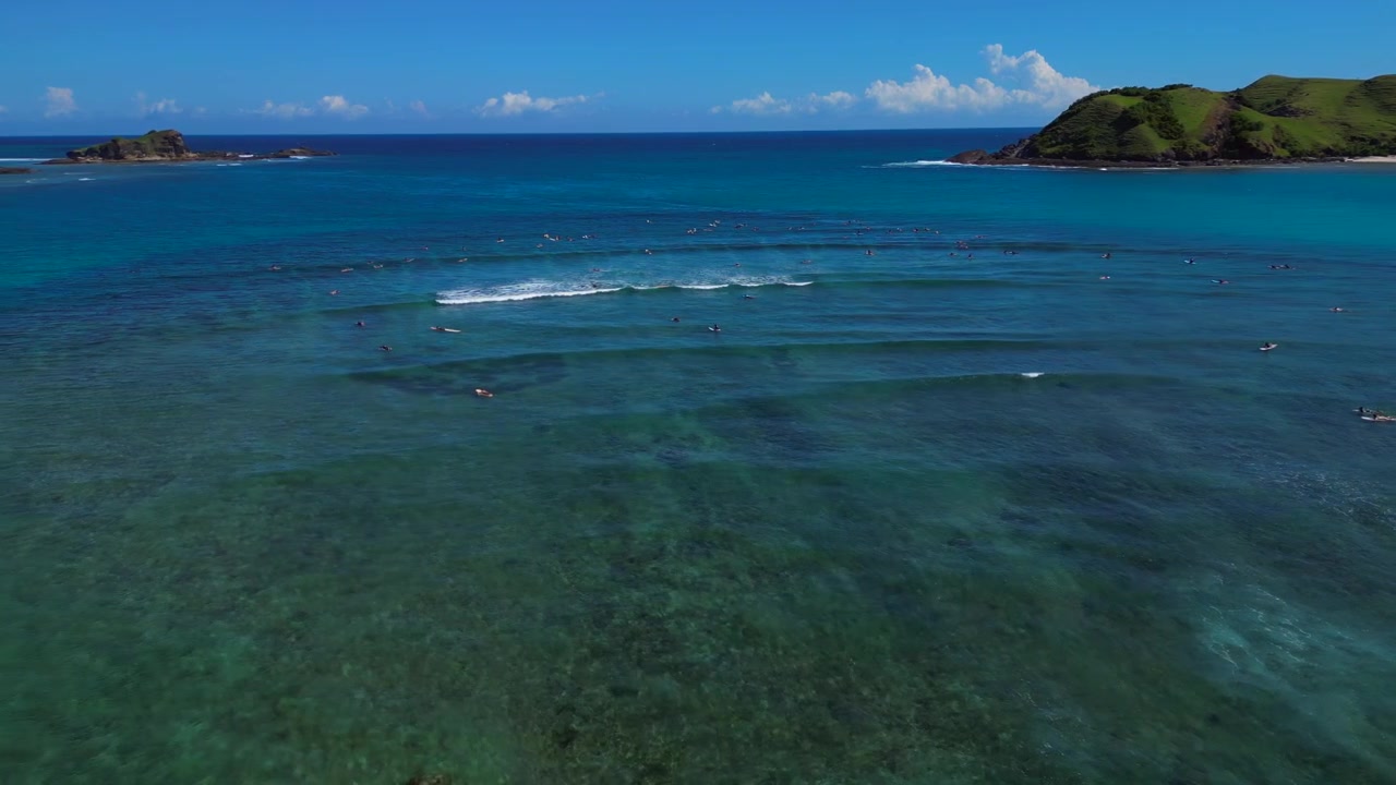 Surfere venter på bølger i det klare blå havet med frodige grønne åser under en lys solrik himmel, som formidler sommer og reise. Fantastisk luftfoto med bred bane, panoramaoversikt fra drone.