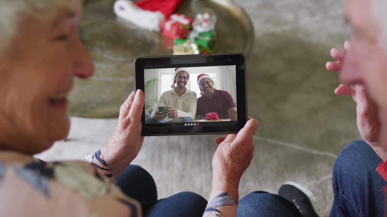 pareja caucásica mayor sonriente usando una tableta para una videollamada de navidad con la familia en la pantalla