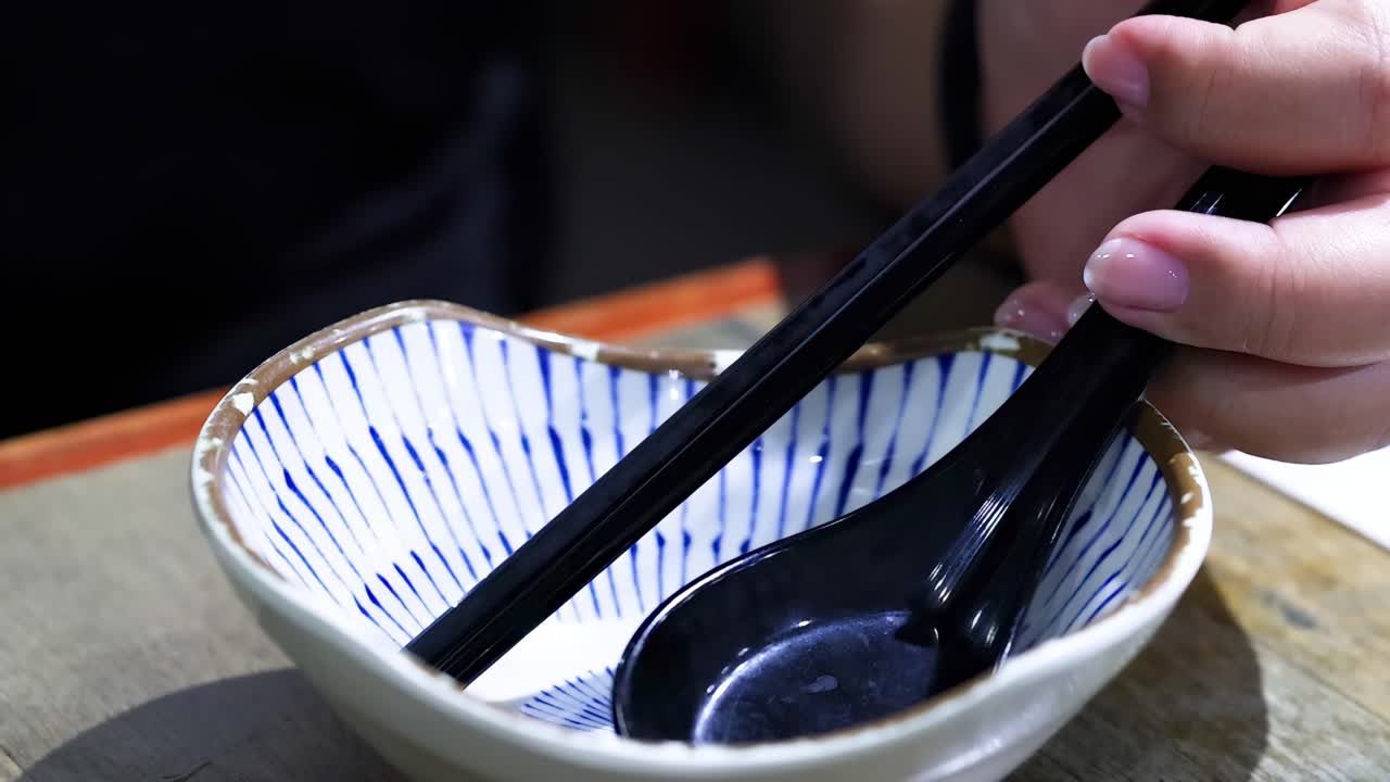 Primo piano di mani che usano bacchette e un cucchiaio in una ciotola di ceramica a strisce con del liquido che viene versato.