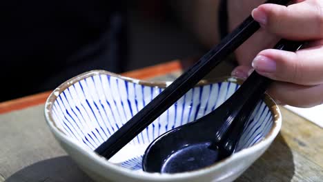 close-up of hands using chopsticks and spoon in a striped ceramic bowl with liquid being poured.