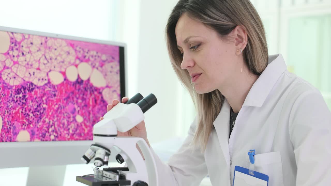 A female scientist examines samples under a microscope in a laboratory, with a magnified view on a computer screen