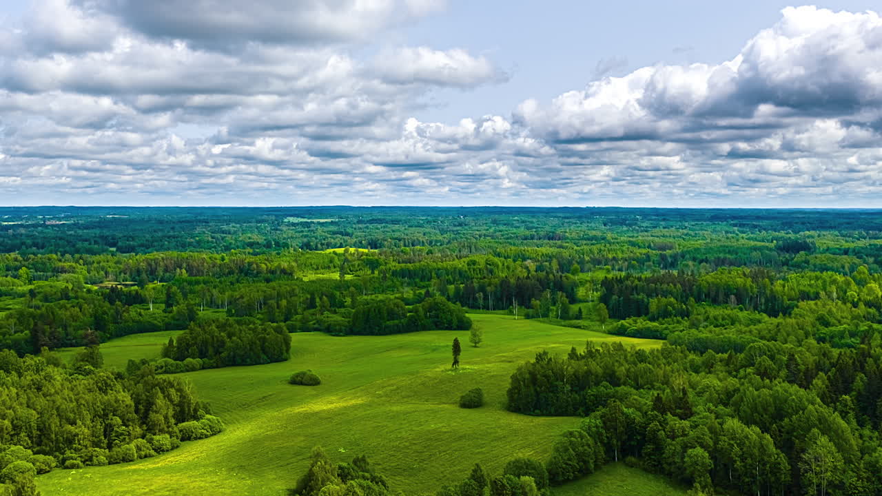 A captivating time-lapse shows dynamic clouds moving across a blue sky, casting shadows over a vast, green Latvian landscape of dense forests interspersed with bright, open grassy clearings