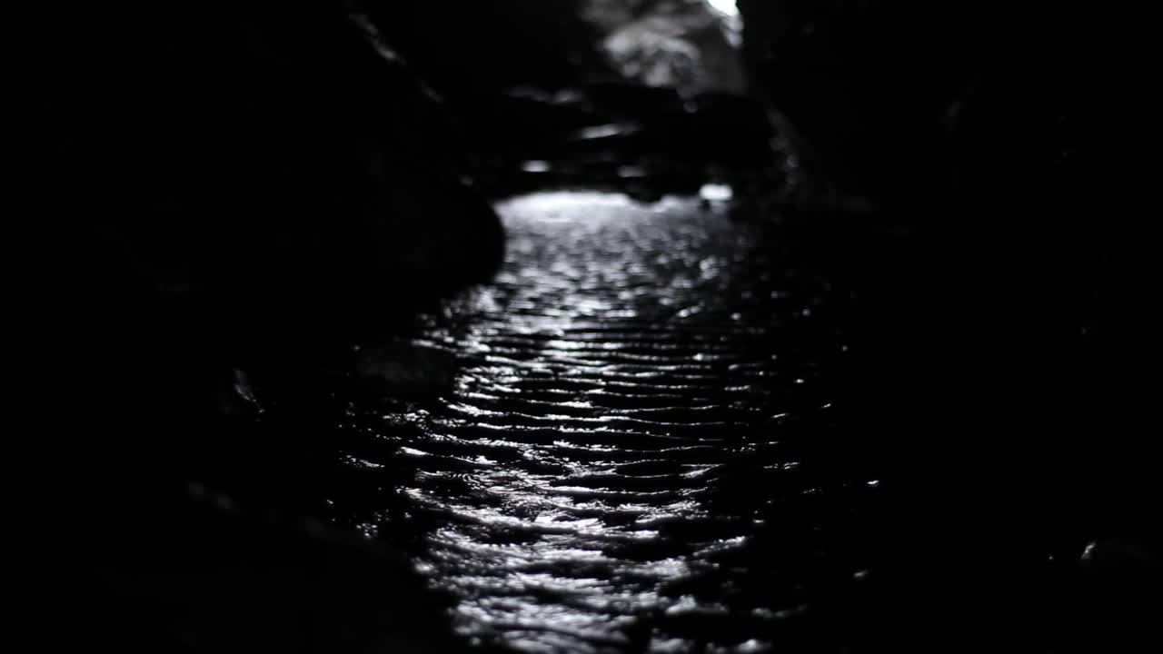 Inside a cave in a mountain ocean brings water at a high tide. It’s dark. Small ripples reflect light coming from entrance on the other end of a cave. It looks mysterious. Cornwall cave. HD