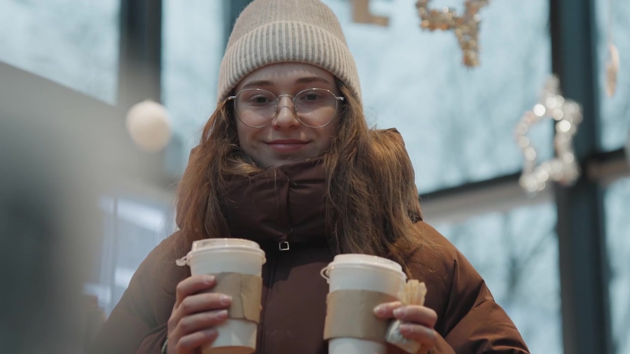 Mulher branca em mercado de inverno segurando cafés, estudante universitária equilibrando dois copos para viagem. Usando gorro de tricô, óculos redondos, casaco acolchoado marrom. Mangas de papelão e tampas em close-up, sob a luz fria da janela, criando um ambiente festivo.