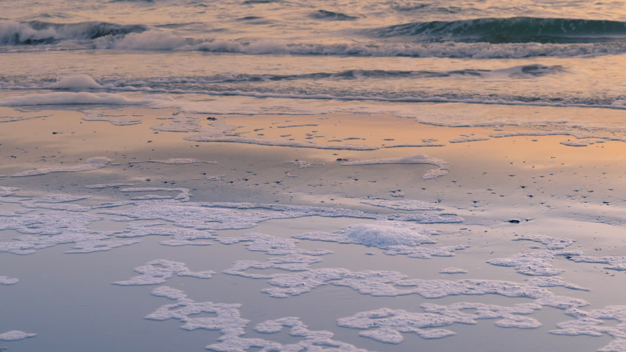 нежные волны промывают песчаный берег при заходе солнца с пеной, разбросанной по пляжу.