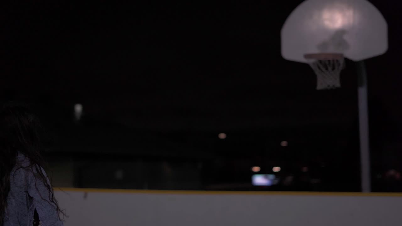 Pige dribler, venter et øjeblik og skyder så bolden om natten på en udendørs basketballbane.