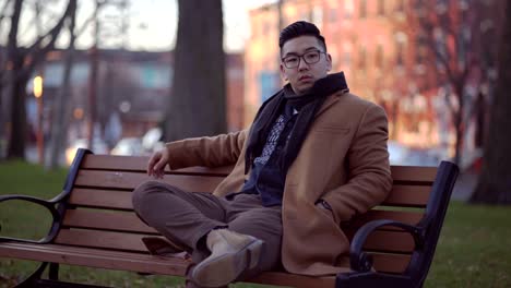 handsome asian korean man wearing in brown coat sitting on the bench chair with glorious trees in the background in new jersey - steady shot