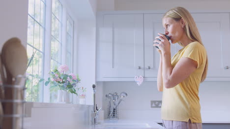 mature woman at home with hot drink standing by kitchen window