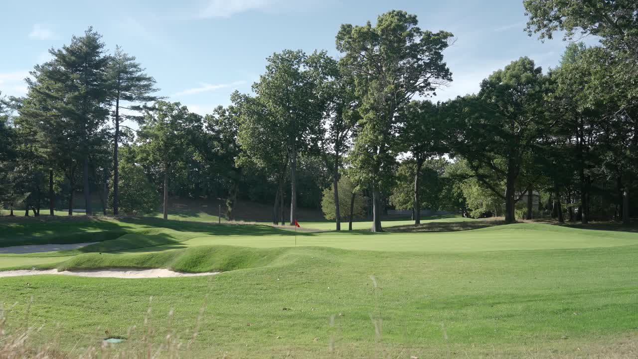 Un putting green di un campo da golf splendidamente mantenuto con una bandiera rossa, circondato da bunker di sabbia e alberi alti in una giornata di sole.