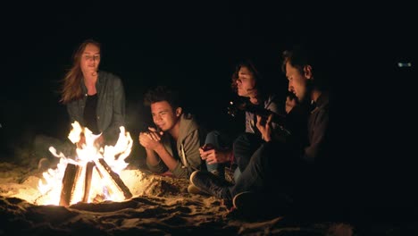 multiracial group of young boys and girls sitting by the bonfire late at night and singing songs and playing guitar