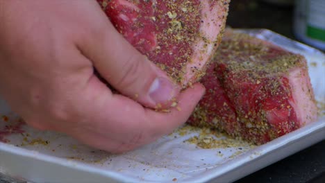 two hands rub spices and salt into a thick raw rib eye steak prepping it to be grilled in slow motion