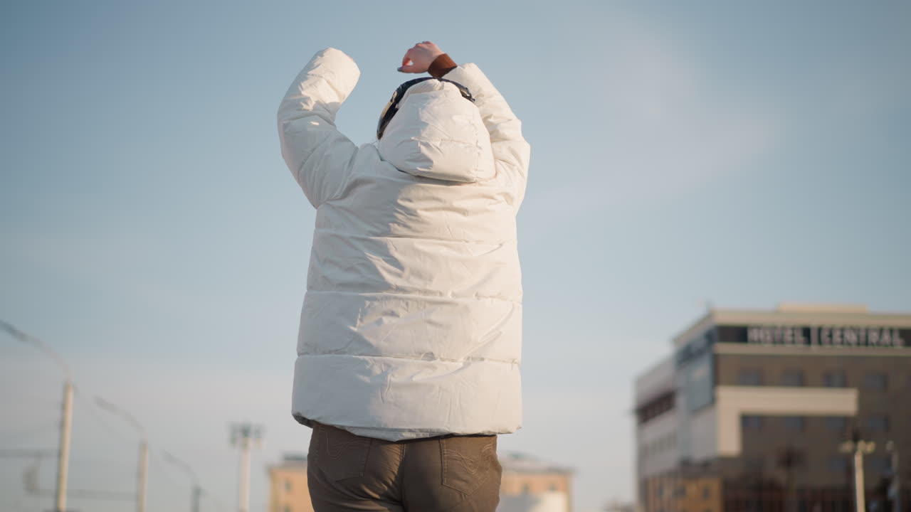 Back view of youth dancing confidently near car park wearing winter jacket and headphones, arms raised in expressive motion while enjoying rhythm under sunlight with hotel sign visible in background