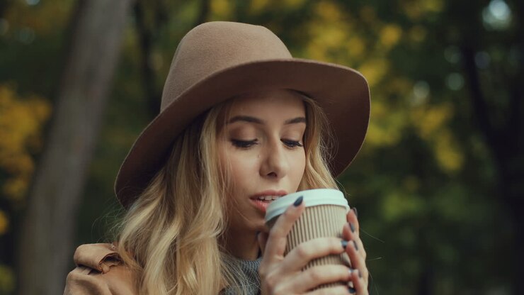 Close-up view of Caucasian young blonde woman in a hat drinking hot coffee sitting on a bench in the park in autumn