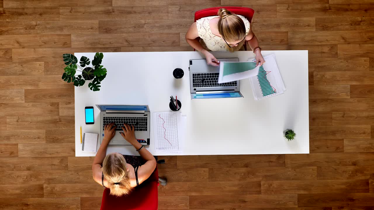 To kolleger gennemgår papirdiagrammer, mens de skriver på bærbare computere, blå skærm på telefonen, scroller, på kontoret