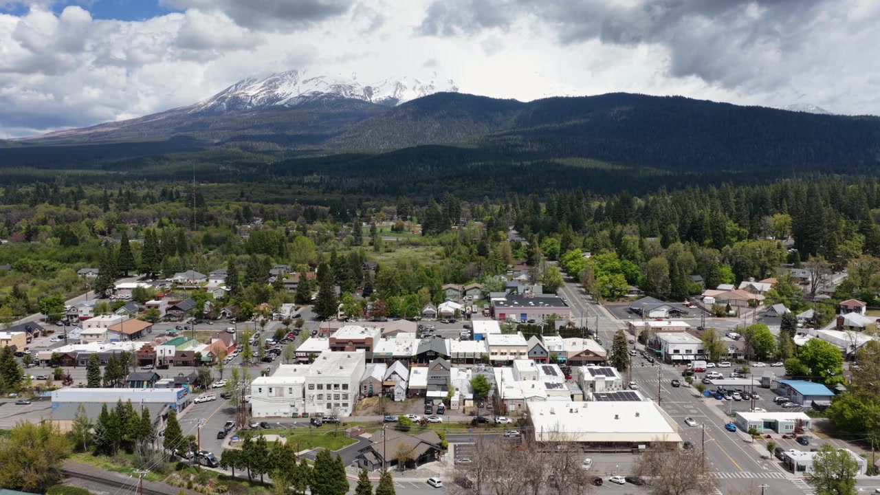 Piccola città con edifici bassi, strade alberate e paesaggio boscoso sotto il Monte Shasta innevato, sotto un cielo nuvoloso e drammatico. Monte Shasta, California, USA. Ampia ripresa aerea all'indietro.