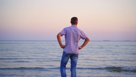 man standing back on the beach and looking at sea in the evening. young man in t-shirt and jeans resting near the sea. handsome man enjoys the nature and breathes fresh air.