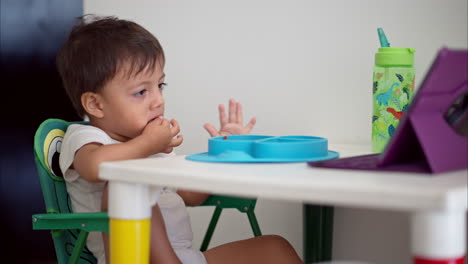 un niñito latino comiendo un panqueque para desayunar mientras ve dibujos animados en una tableta morada sentado en una mesa blanca
