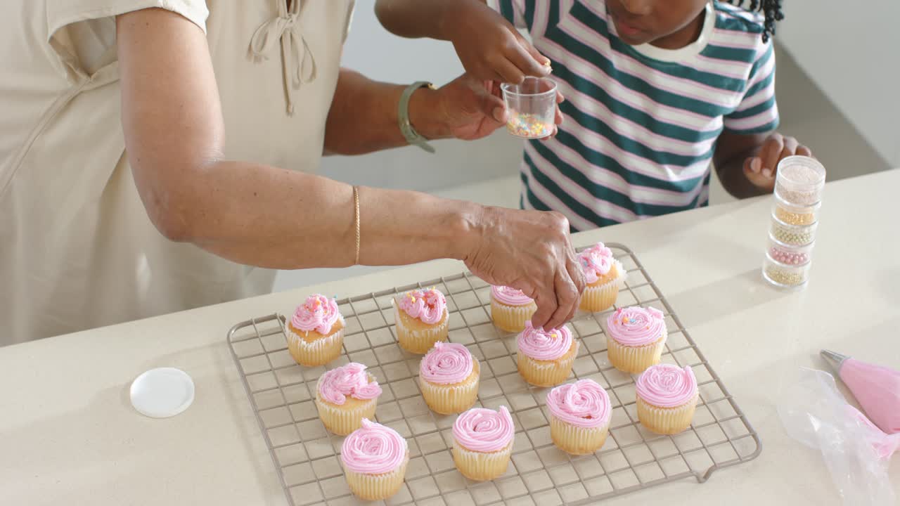 Mulher idosa afro-americana ensinando uma menina a decorar cupcakes no balcão, oferecendo um copinho de granulado.