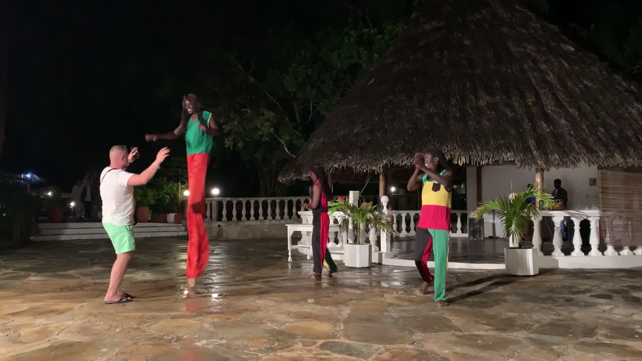 European - caucasian tourist dancing with stilted black dancers at Kenyan hotel at Diani Beach. White tourist enjoying black dance performance, reminiscent of colonialism.