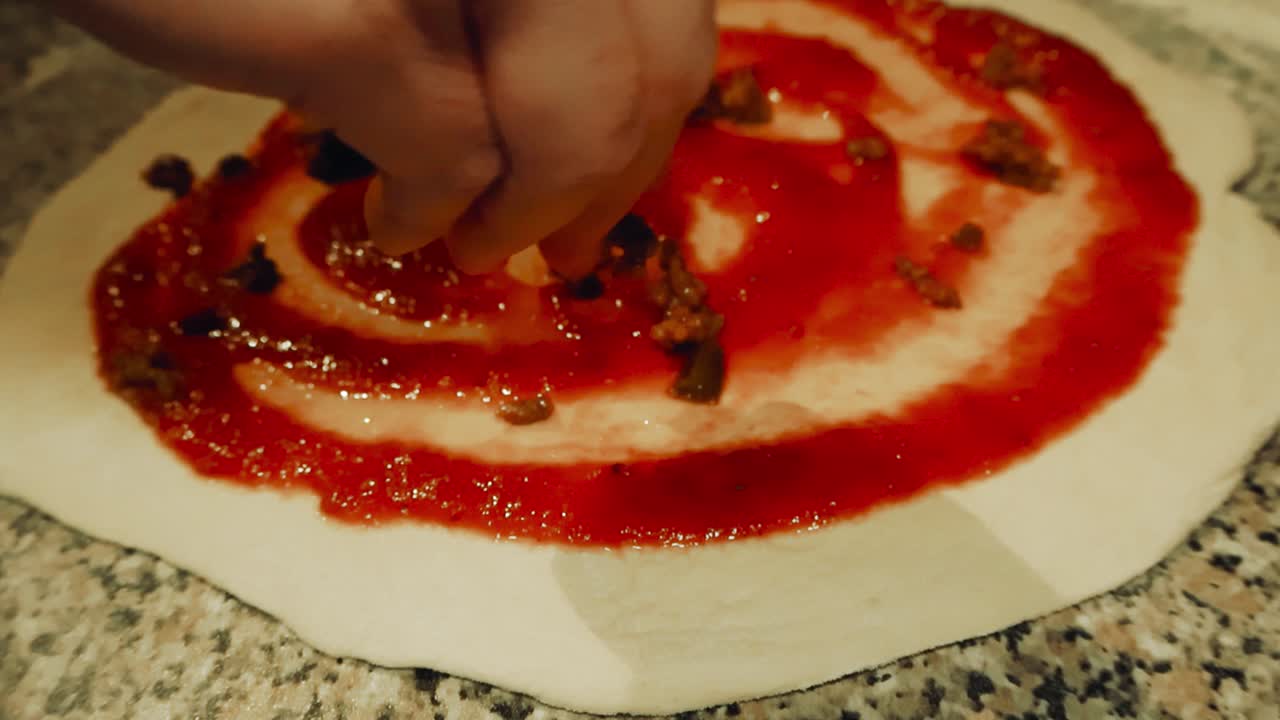 Slow motion video of a hand spreading and topping raw pizza dough with tomato sauce on top with cooked minced meat at a home kitchen where white flour is around on the table. Shallow depth of field