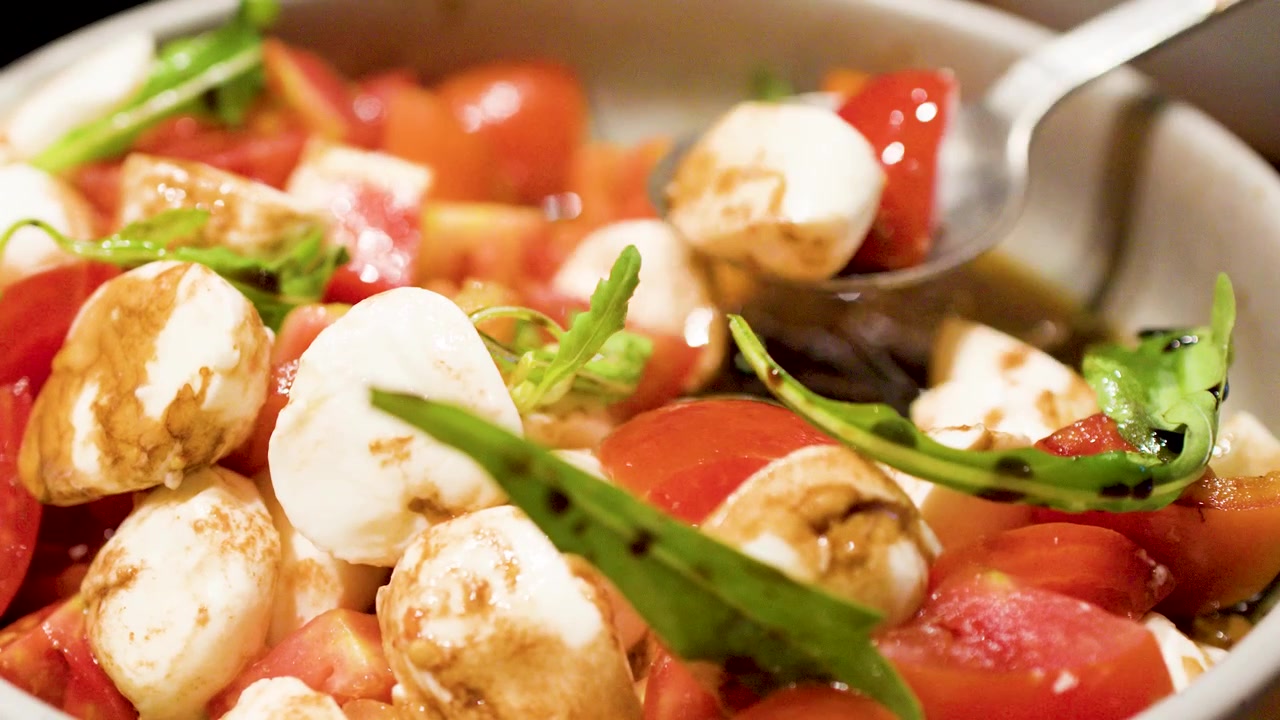 Un cucchiaio di metallo raccoglie palline di mozzarella fresca e pomodori da una ciotola con glassa balsamica.
