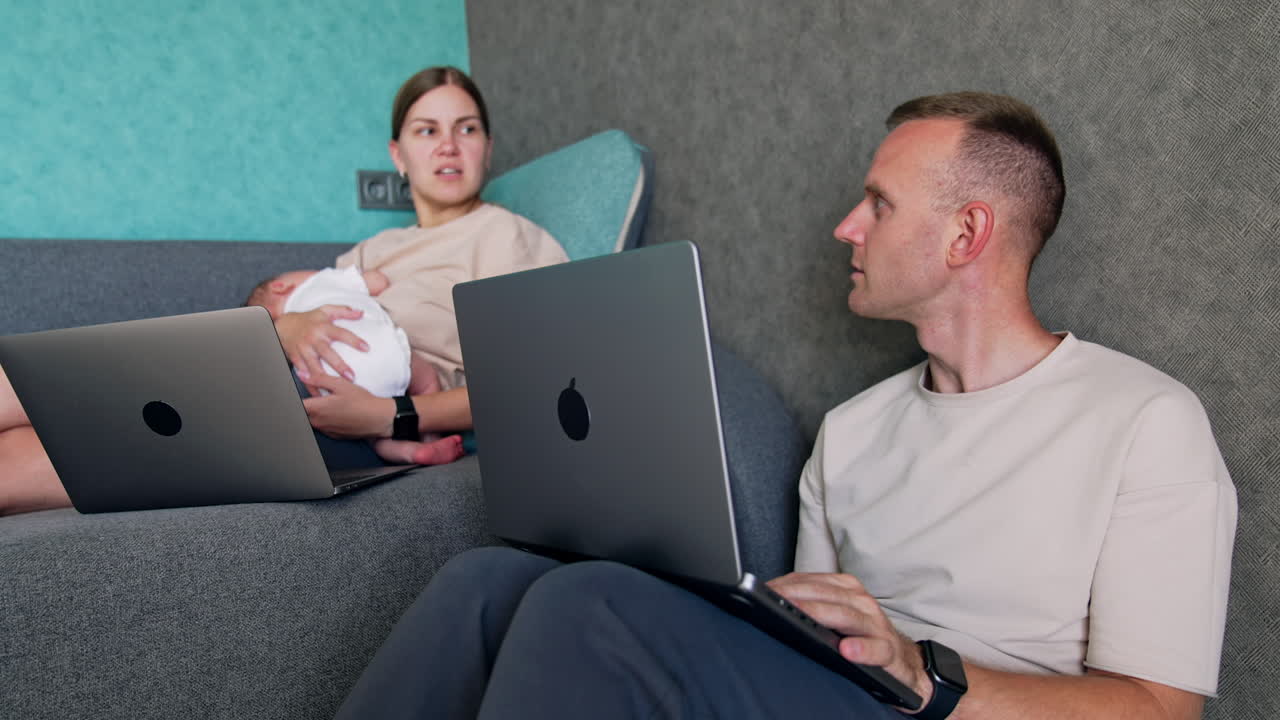 Busy modern parents of newborn working at home. Caucasian couple nurse their baby and work via internet.