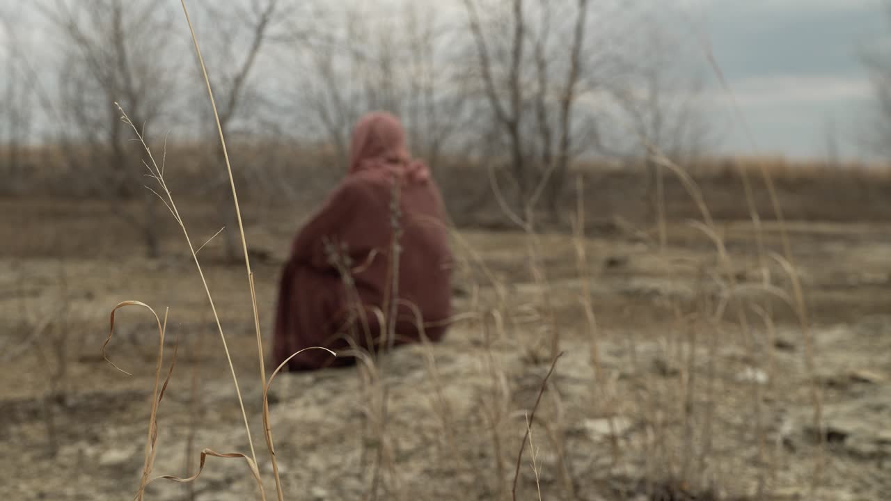 A middle eastern man wearing robes, tunic and hood sits on the rocky ground in the distance surrounded by trees without leaves on a cloudy and overcast day in slow motion.