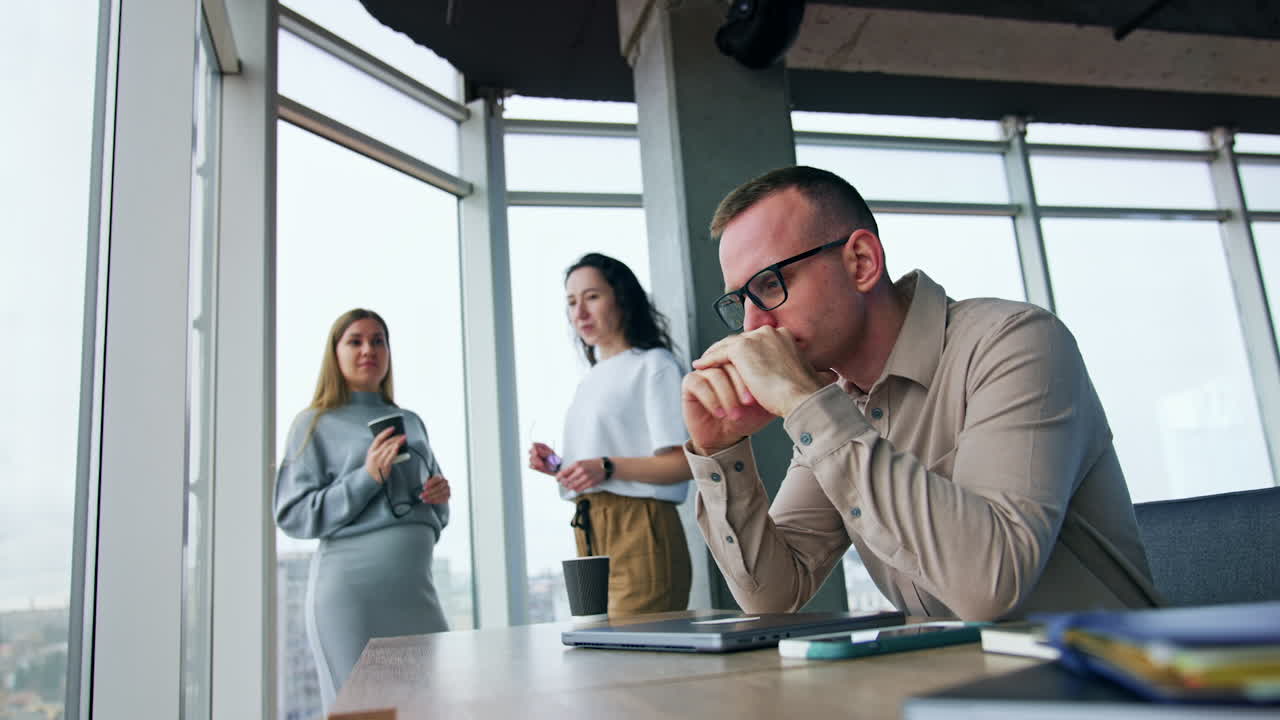 Stressed tired male employee sits at desk. Man takes off his eyeglasses touching his head. Female colleagues talk at backdrop. Low angle view.