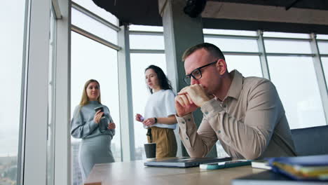 stressed tired male employee sits at desk. man takes off his eyeglasses touching his head. female colleagues talk at backdrop. low angle view.