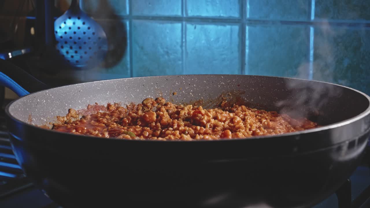 Ragù alla bolognese vegetariano che sobbolle e rilascia vapore in una padella nera su un fornello, con cucchiai da cucina sullo sfondo.