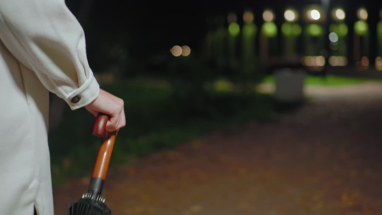 Close up partial view of lady walking at night holding umbrella by wooden handle, wearing light coat, with blurry bokeh car lights in background and dim streetlight illuminating park pathway
