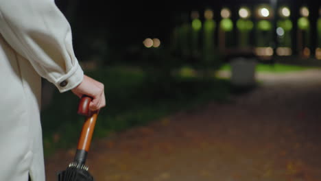 close up partial view of lady walking at night holding umbrella by wooden handle, wearing light coat, with blurry bokeh car lights in background and dim streetlight illuminating park pathway