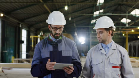 Two-engineers-wearing-helmets-holding-tablet-while-talking-and-walking-in-a-factory