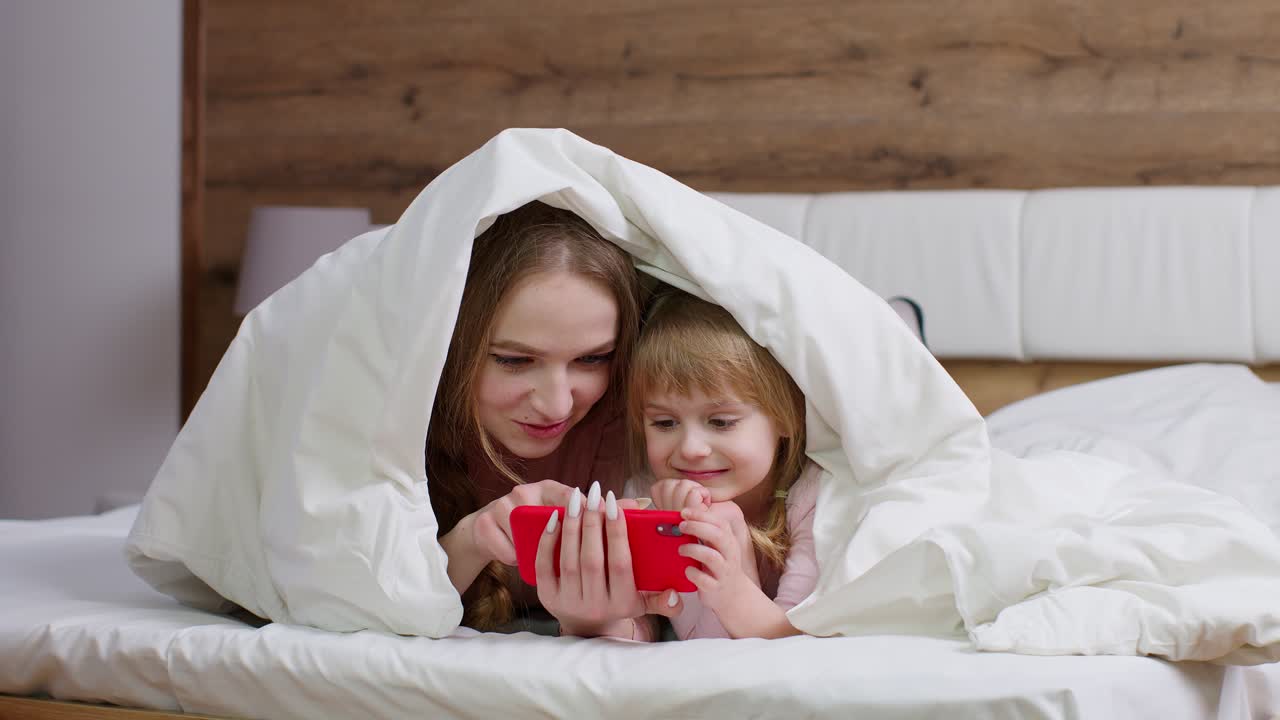 Madre sdraiata a letto sotto una coperta con la figlia bambina con il cellulare che guarda un cartone animato